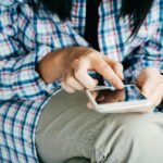 Woman's hands using a smartphone, wearing plaid shirt, indoors. Close-up shot highlighting touch interaction.